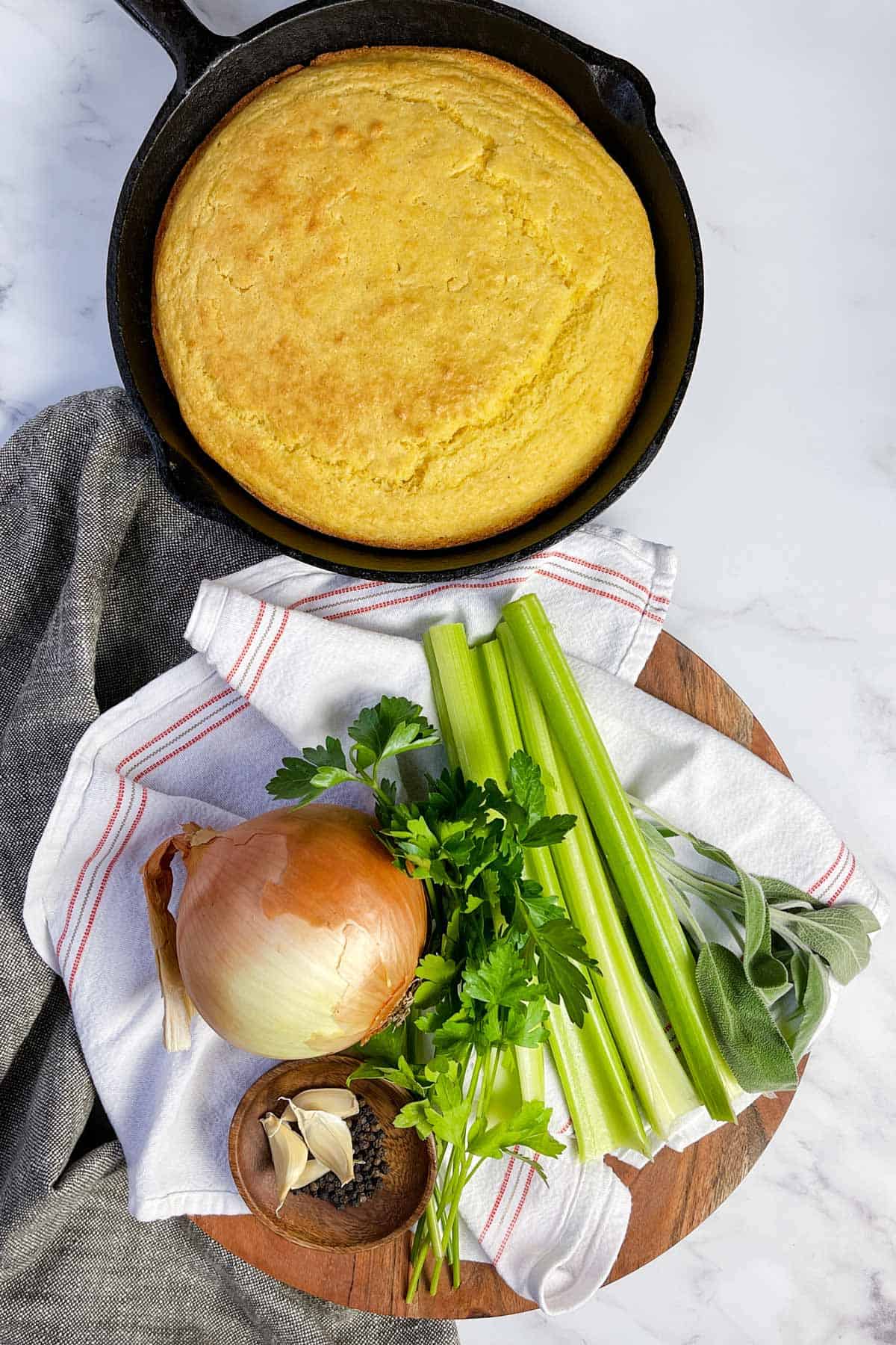 A skillet of cornbread dressing rests on a marble surface, surrounded by an onion, celery stalks, garlic cloves, fresh herbs, and a small bowl of peppercorns on a wooden board with a striped cloth.