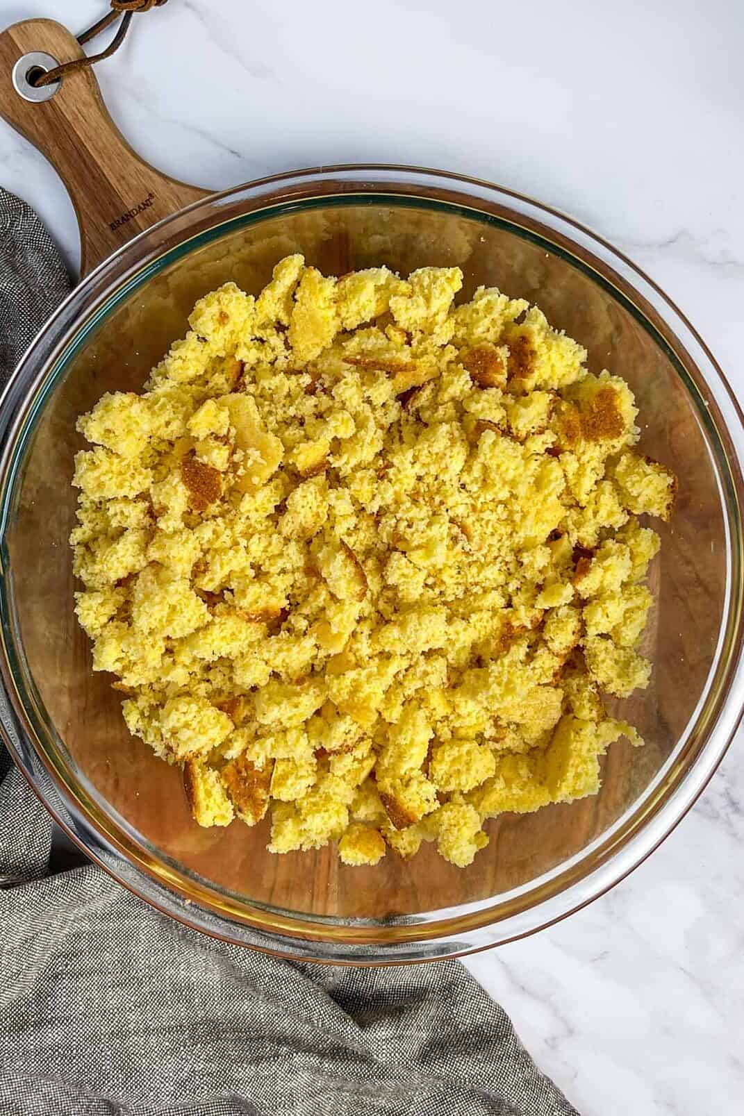 A glass bowl filled with crumbled cornbread, perfect for making delicious cornbread dressing, sits on a wooden cutting board. A gray cloth napkin is partially visible next to the board, while the background showcases a sleek marble surface.