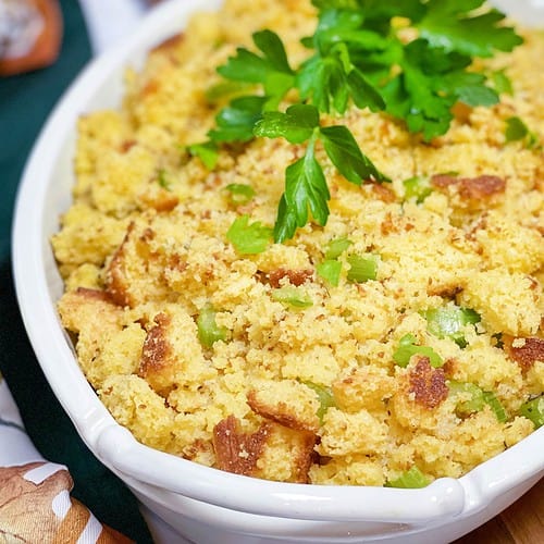 A white dish filled with cornbread dressing garnished with fresh parsley sits invitingly. The dressing boasts a golden-brown, crumbly texture contrasted by green celery pieces. A wooden spoon and a green cloth napkin complete the picturesque setting.