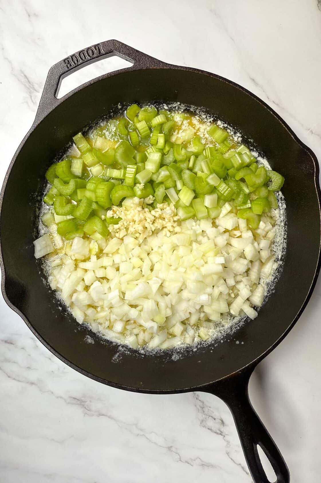 A black cast-iron skillet on a marble surface contains chopped celery, minced garlic, and diced onion being sautéed in melted butter. The ingredients are visibly separated in the pan.