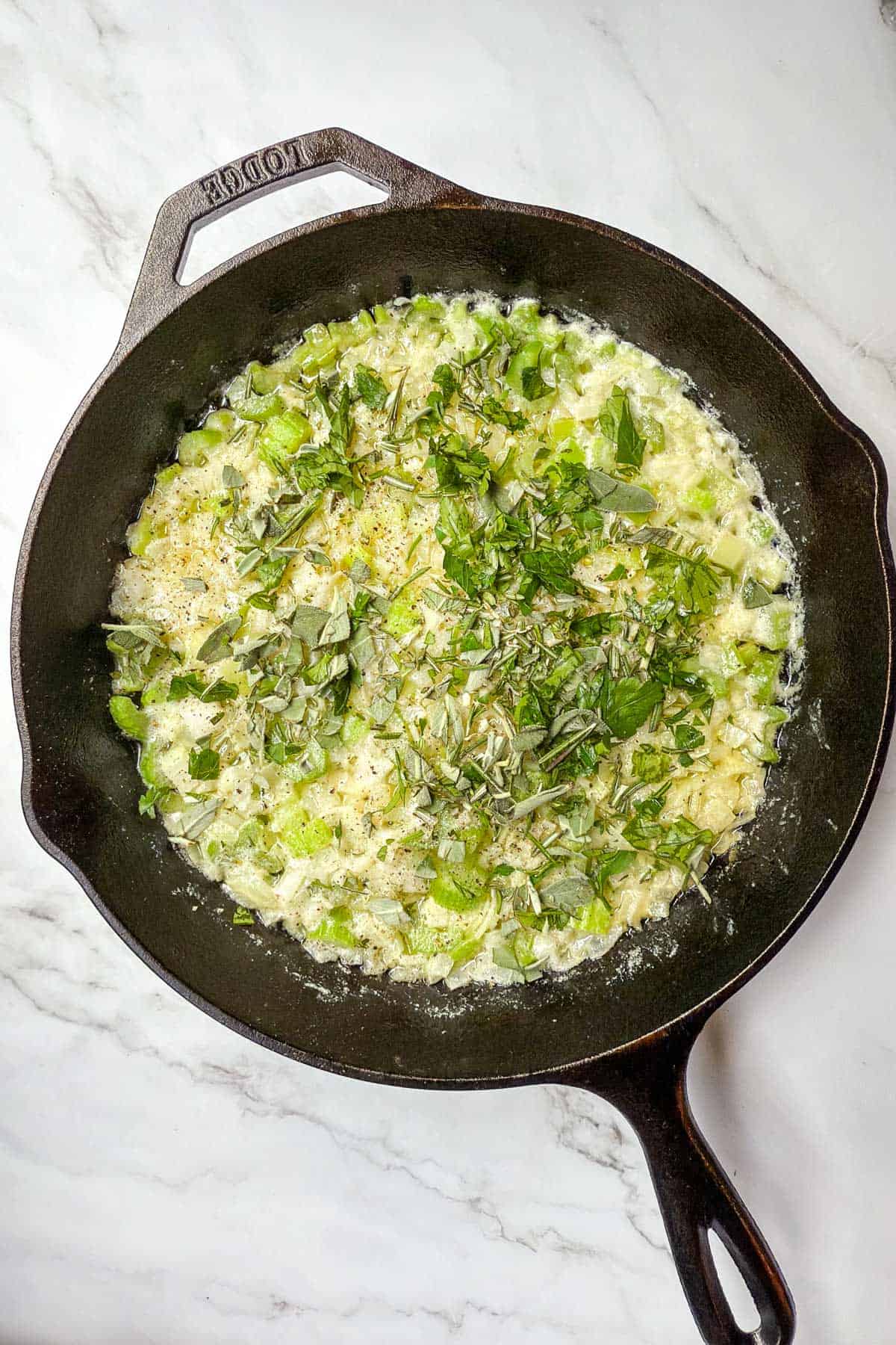 A cast iron skillet filled with a mixture of chopped onion and celery topped with fresh herbs is placed on a marble countertop.