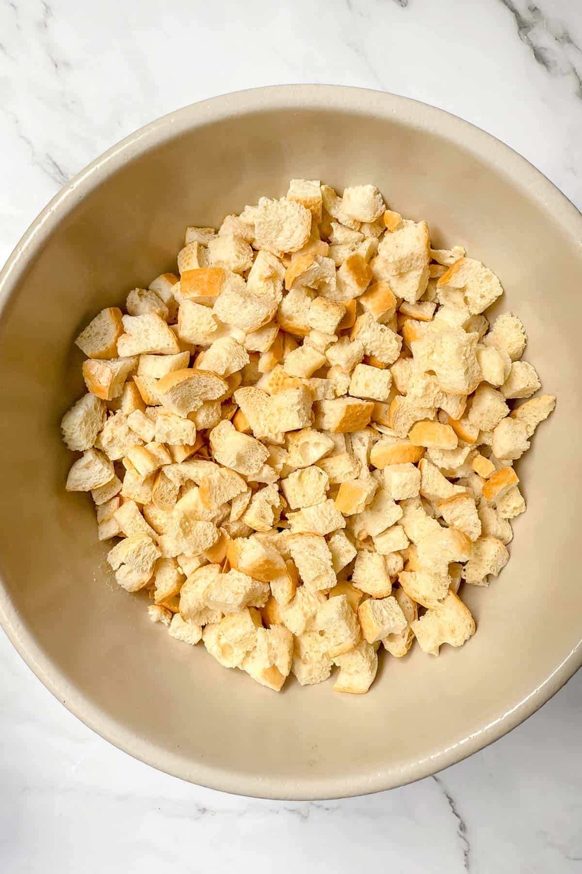 A beige bowl filled with small toasted cubes of bread sits on a white marble surface. The bread cubes are light golden brown in color and appear crispy.