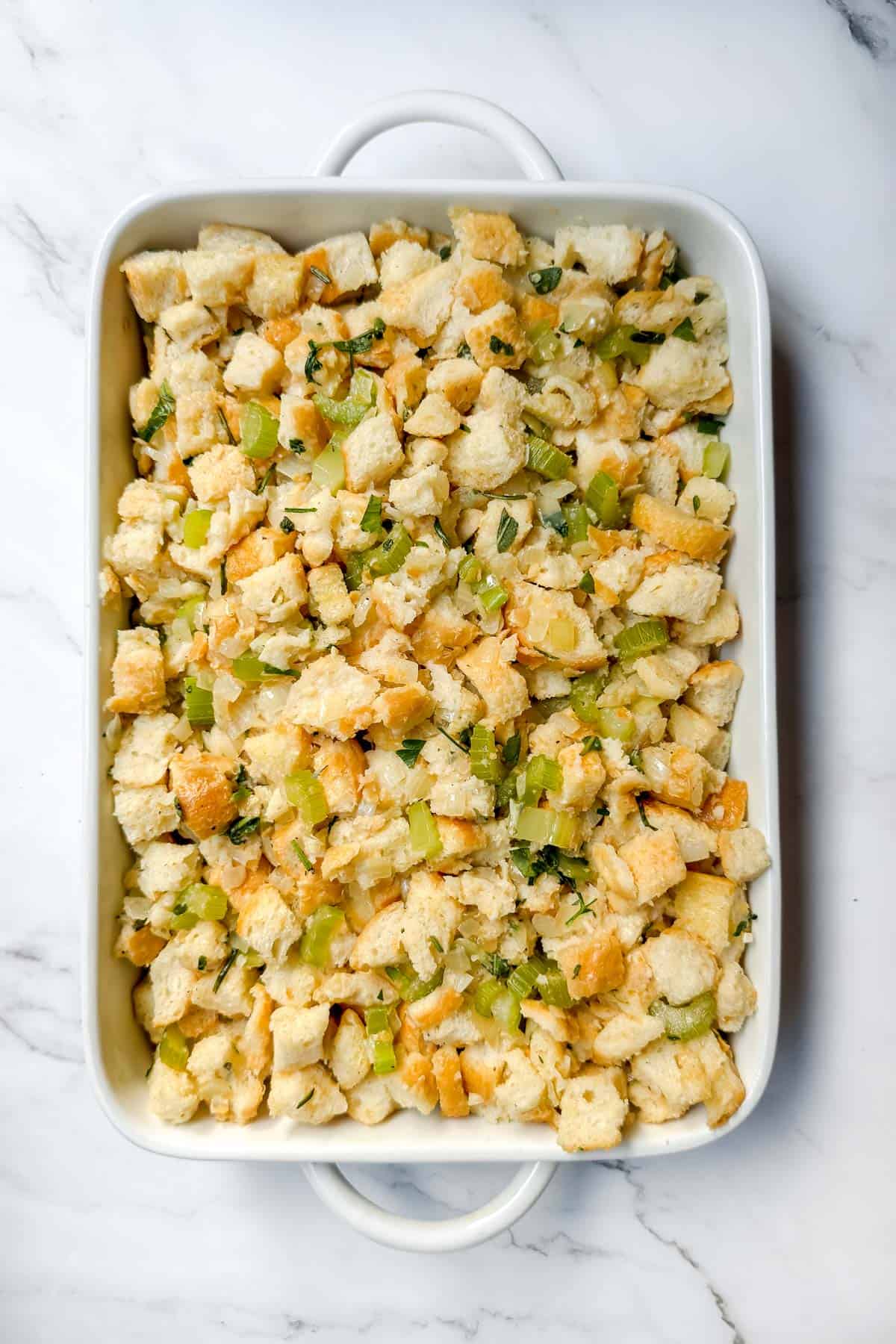 A white baking dish filled with bread stuffing, featuring chunks of bread, chopped celery, onions, and herbs. The mixture is ready for baking, set on a marble countertop.