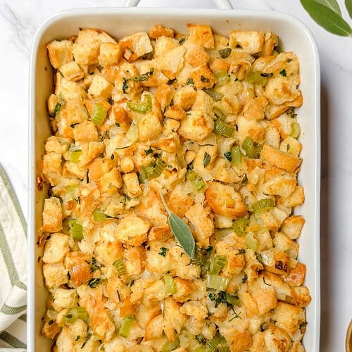 A white casserole dish filled with golden-brown stuffing, featuring cubed bread, celery, and herbs. A fresh sage leaf garnishes the top. A striped kitchen towel is placed beside the dish on a marble surface.