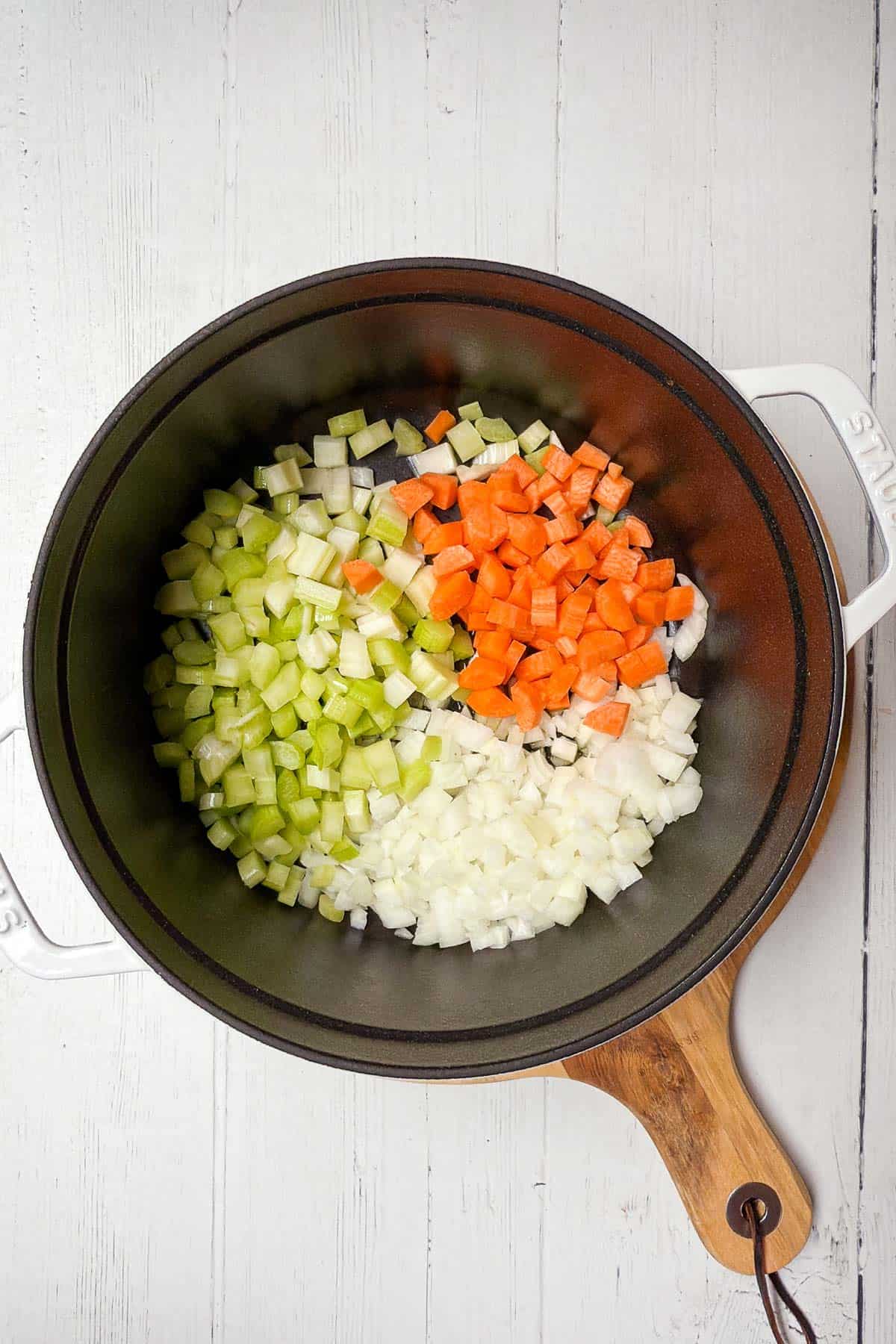 A pot on a wooden board containing diced vegetables, including green celery, orange carrots, and white onions, arranged in separate sections. The pot is placed on a white wooden surface.