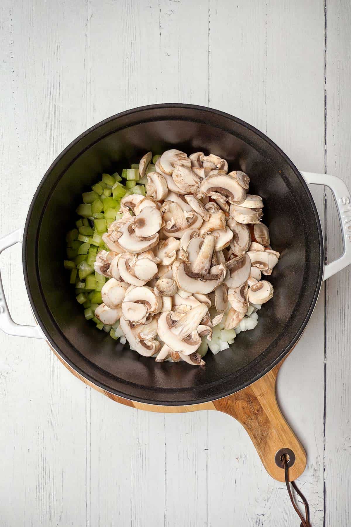 A top-down view of a black pot on a wooden board. Inside are chopped celery, sliced mushrooms, and diced onions. The pot's handles are white, and the scene is set against a white wooden background.