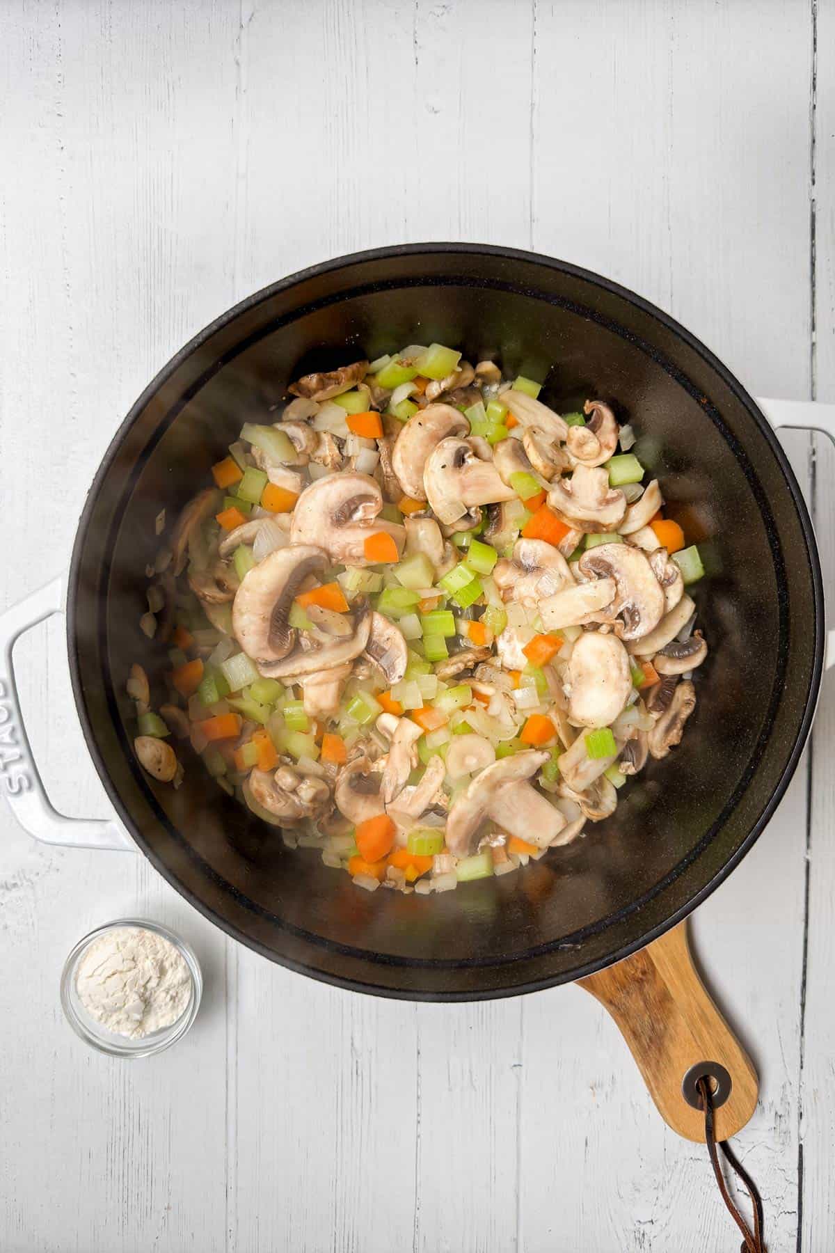 A black pan on a wooden handle contains sautéed mushrooms with chopped celery, carrots, and onions. The mixture is on a white background, and there's a small bowl of flour beside the pan. Steam is visible rising from the pan.