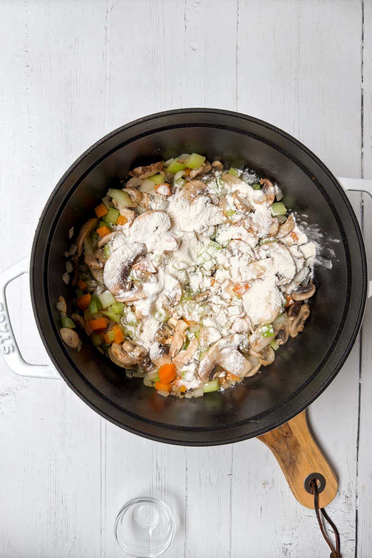 A black pot with chopped vegetables including carrots, celery, and mushrooms, topped with a dusting of flour, sits on a white wooden surface. A wooden-handled, lid-less pot is positioned diagonally.