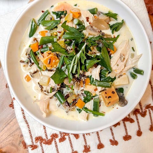 A white bowl filled with a creamy turkey wild rice soup. Garnished with fresh herbs, the bowl is placed on a wooden board with a white and brown embroidered cloth.