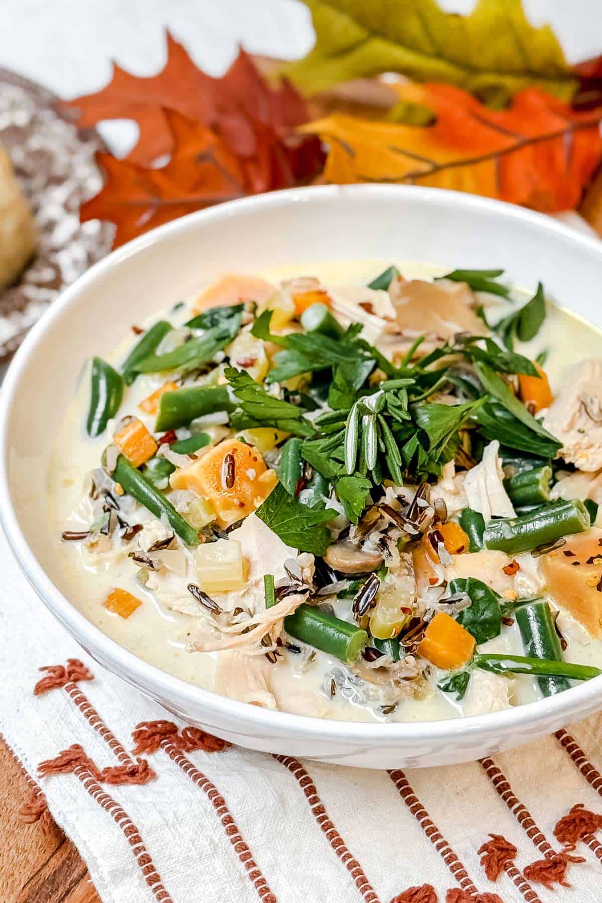 A bowl of creamy soup with shredded turkey, green beans and wild rice. The dish is garnished with fresh parsley and chili flakes, set on a white tablecloth. Autumn leaves in the background suggest a fall theme.